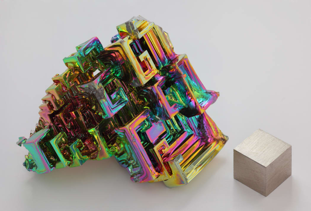 The chemical element bismuth as a synthetic crystal. The iridescent surface is a very thin layer of oxidation. Beside it is a high purity (99.99 %) 1 cm3 cube of bismuth for comparison.