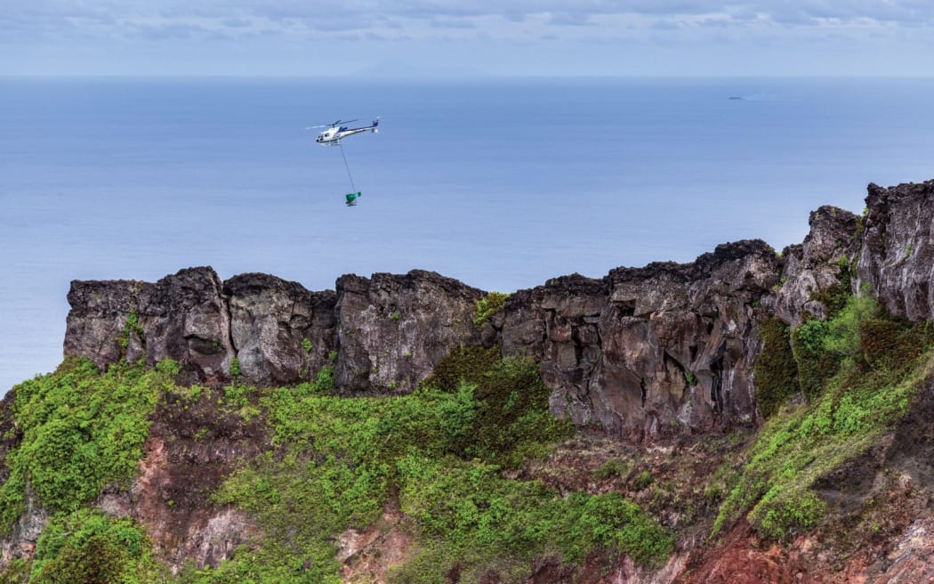 A helicopter with a bucket attached via rope swinging underneath it flies along a rocky, sharp ridgeline with bright green vegetation on its slopes. The ocean is visible in the background, beneath a cloudy sky. The hazy outlines of other islands are visible on the horizon.
