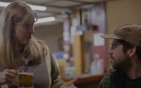 Brynley Stent, a white woman with blonde hair, holds a beer and speaks with a seated dark-haired man in cap.