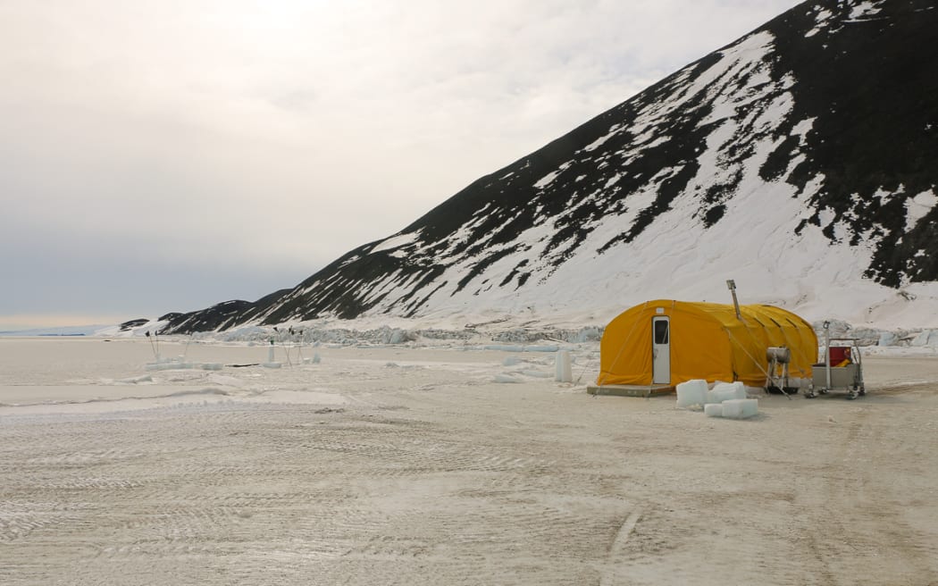 A landscape photo of a large yellow tent on an expanse of ice with vehicle tracks across it, in front of a rocky mountain with snow. The sky is overcast.