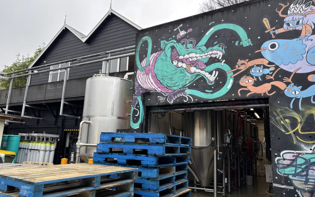 A black warehouse building with a colourful surreal mural of strange creatures on its facade. In front of the entrance, there are wooden pallets stacked high and a large metal drum.