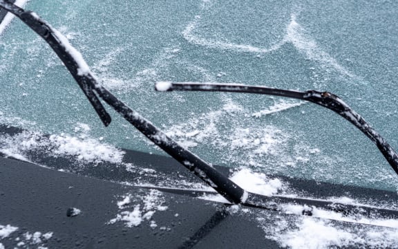 Windshield of car is covered with light snow and wipers are raised from glass.