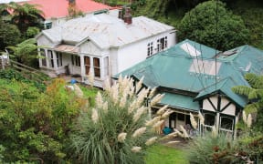 Two old villas in a gully. On the left, a white house with rusted roof panels. On the right, a cream house with a green roof with miscoloured replacement panels. Both look dark and cold.