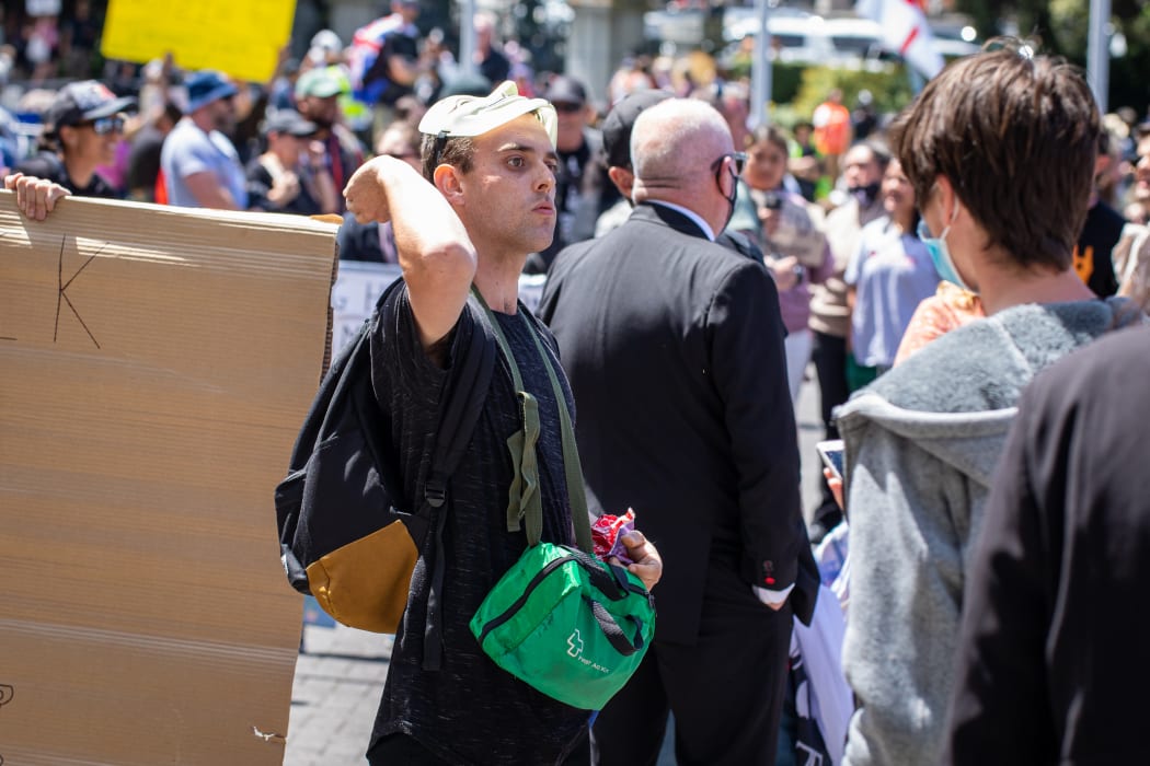 A protestor in a V-for-Vendetta mask takes idle potshots at media and police with his supply of farmbake cookies.