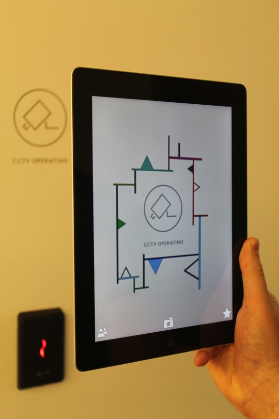 An image of a tablet held in front of a CCTV sign revealing the artwork digitally attached to it.