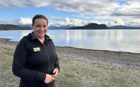 A woman wearing a fleece with a 'Project Tongariro' badge. She is standing on a lakeshore and smiling beneath a cloudy blue sky.