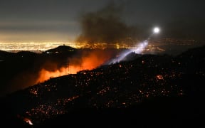 The light of a fire fighting helicopter illuminates a smouldering hillside as the Palisades fire grows near the Mandeville Canyon neighborhood and Encino, California, on January 11, 2025. The Palisades Fire, the largest of the Los Angeles fires, spread toward previously untouched neighborhoods January 11, forcing new evacuations and dimming hopes that the disaster was coming under control. Across the city, at least 11 people have died as multiple fires have ripped through residential areas since January 7, razing thousands of homes in destruction that US President Joe Biden likened to a "war scene." (Photo by Patrick T. Fallon / AFP)