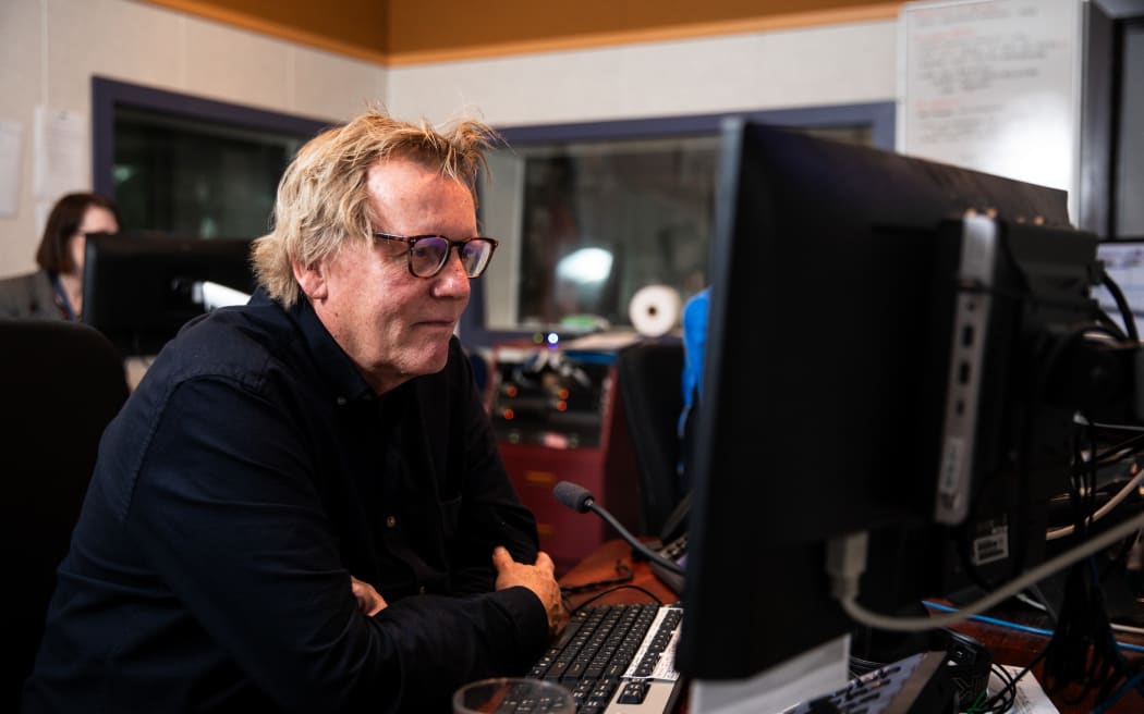 A man sits a a desk with a computer screen in front of him. He is wearing a dark shirt and glasses.