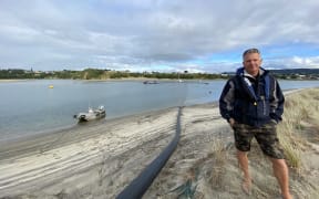 Mangawhai Harbour Restoration Society dredge operator Mark Vercoe on the sandspit where the barge in the background pumps sand along the black pipe beside him, to keep the harbour open and build up the protective sand dune barrier known as a bund wall that protects the sandspit from harbourside-generated erosion.