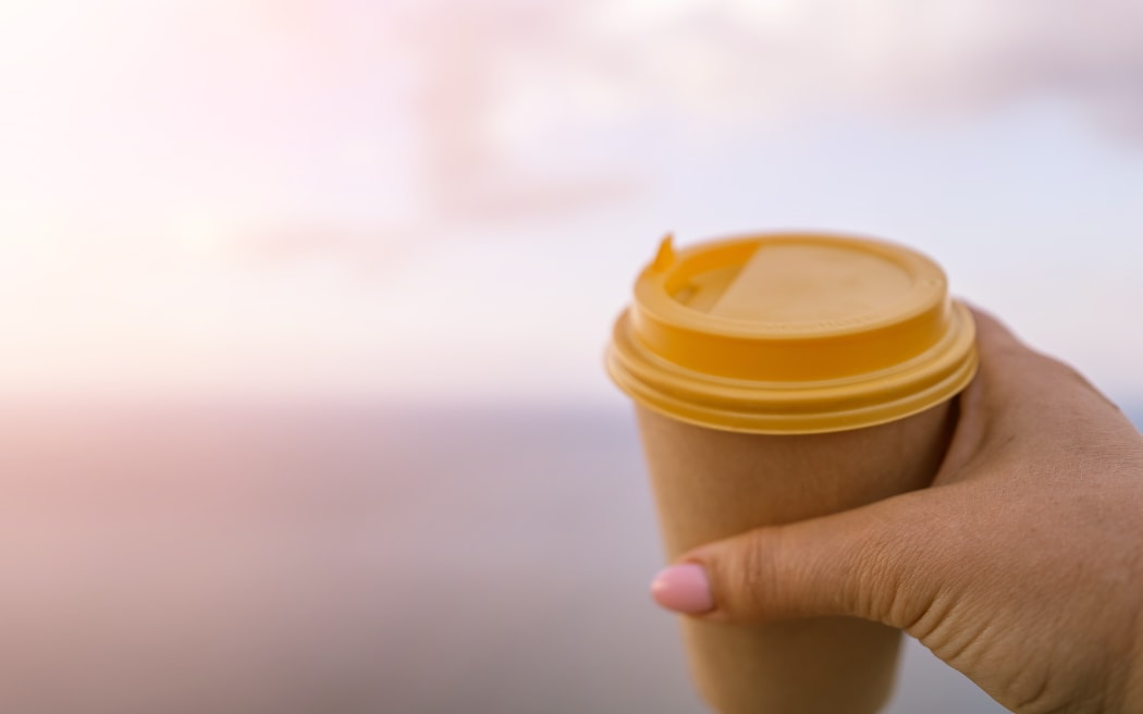 Hand holding Yellow cup with lid, coffee against a backdrop of a blue sky and sea. Illustrating cup and beverage.