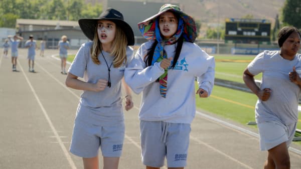 Harper (Julia Butters) and Lily (Sophia Hammons) running on a track, wearing wide-brimmed sunhats and sunscreen in the Disney film Freakier Friday.