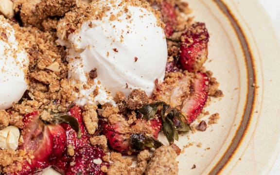 A close-up of a beige plate on a white table. The plate is strewn with crumble, chunks of strawberry, and three lumps of ice cream.