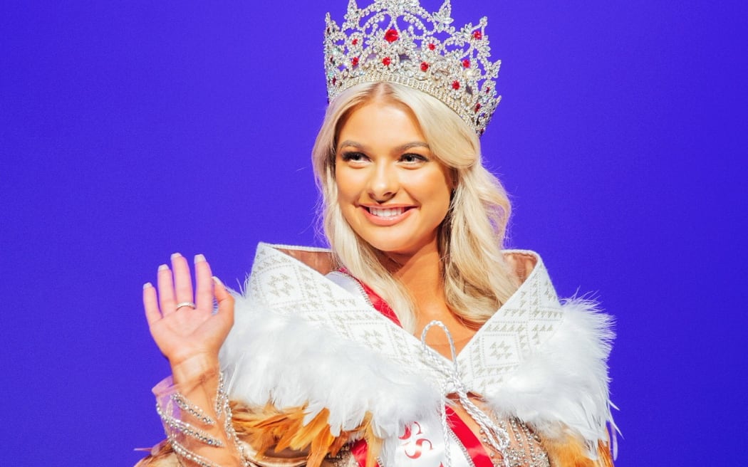 Georgia waves to a crowd. She is wearing a large silver and red crown, a sash saying 'MISS NEW ZEALAND', and a cloak of feathers.