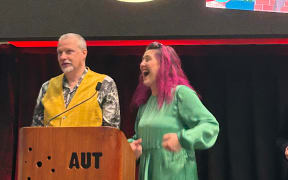 Louise & Gareth Ward are onstage collecting an award at the 2025 'Book industry awards'. Gareth is wearing a waistcoat and looking coy. Louise has her trademark pink hair. She is wearing a green dress and looks very excited. The cover of their book is projected on the stage behind them.