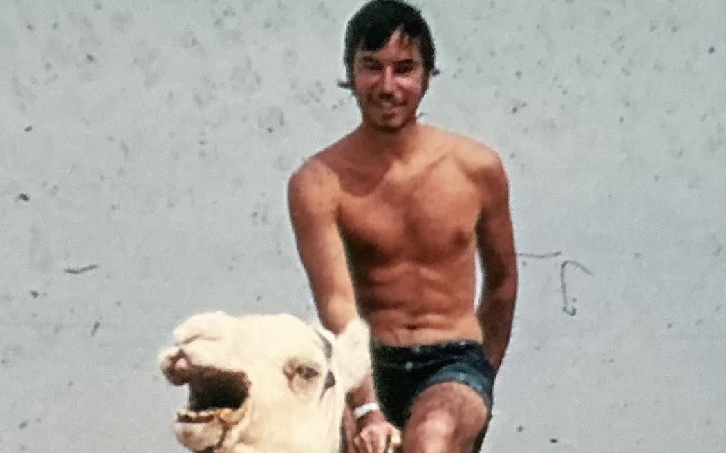 Chris Watson is riding a camel through the Sahara desert. He is shirtless. The sand behind him is very flat.