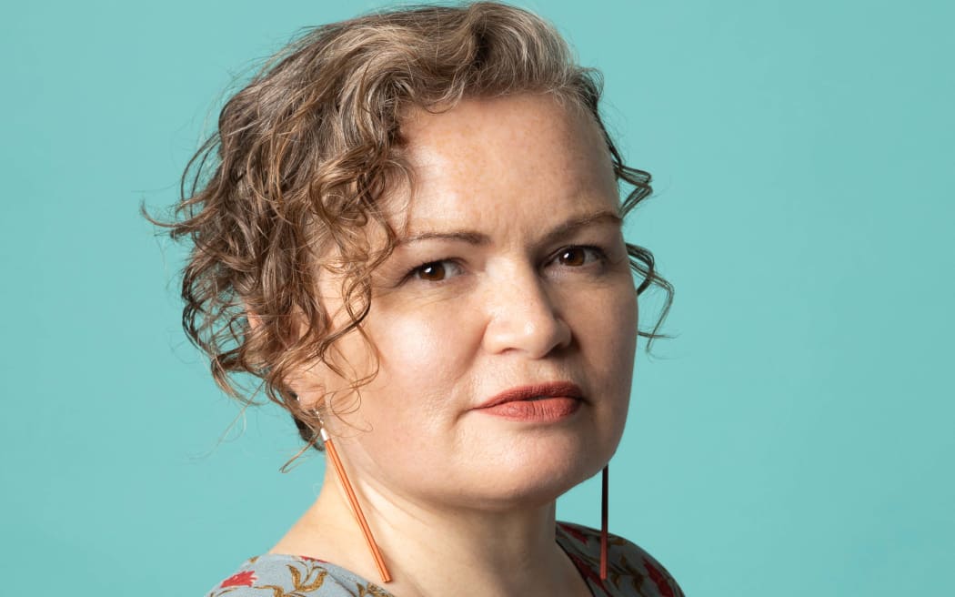 Tina stands in front of a blue background. She has cheek length wavy hair falling dow the left hand side of her face. She is wearing earrings which are like long orange, dangly sticks.