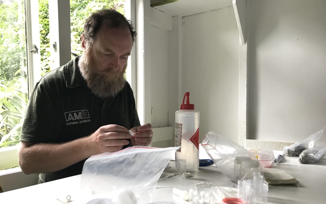 A man with a beard sits at a white bench with plastic ziploc bags and vials in front of him.