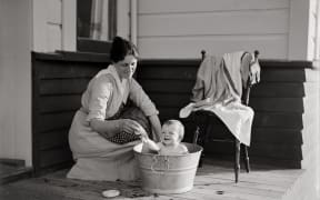 Maud bathes her baby Nancy in a metal tub on the deck of the house. They both smile.