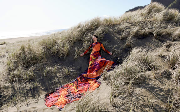 Image of model reclining in sand dunes wearing long brightly coloured (orange, black, yellow, pink) gown with train. Zheyi Ruan entry for 2023 iD Dunedin Emerging Designer Awards.