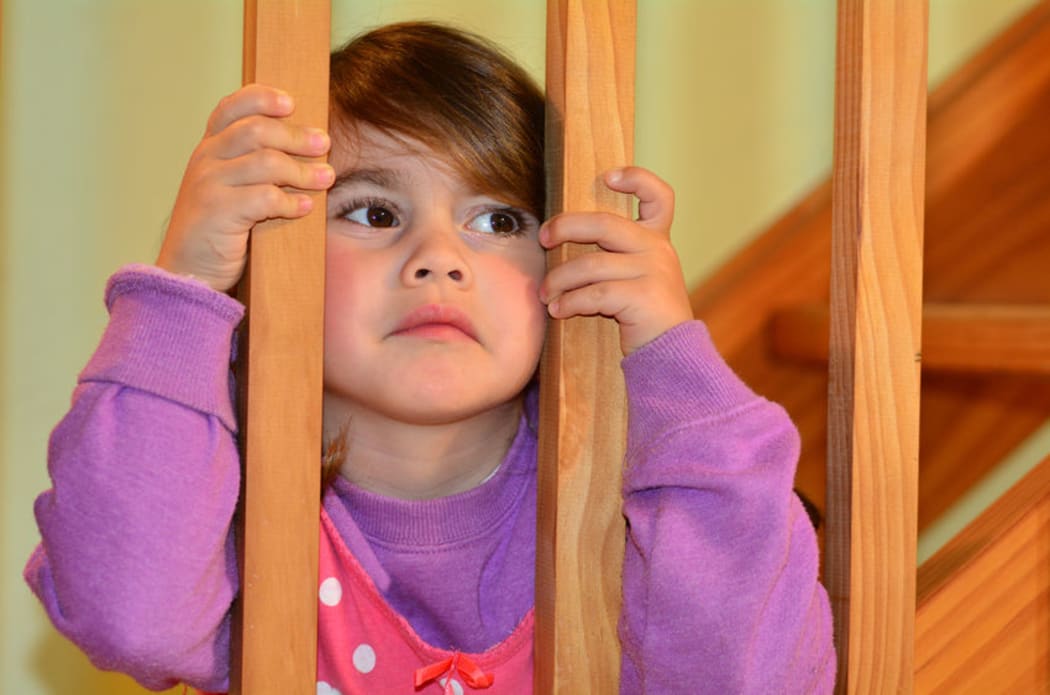 A photo of a young, anxious looking girl peering through the bars of the stairs