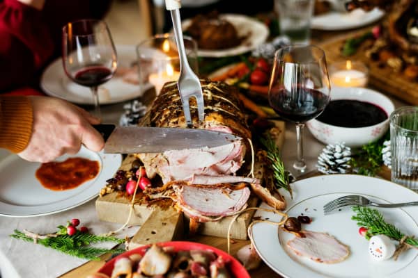 A person slicing roasted ham on a dinner table full of food plates and wine.