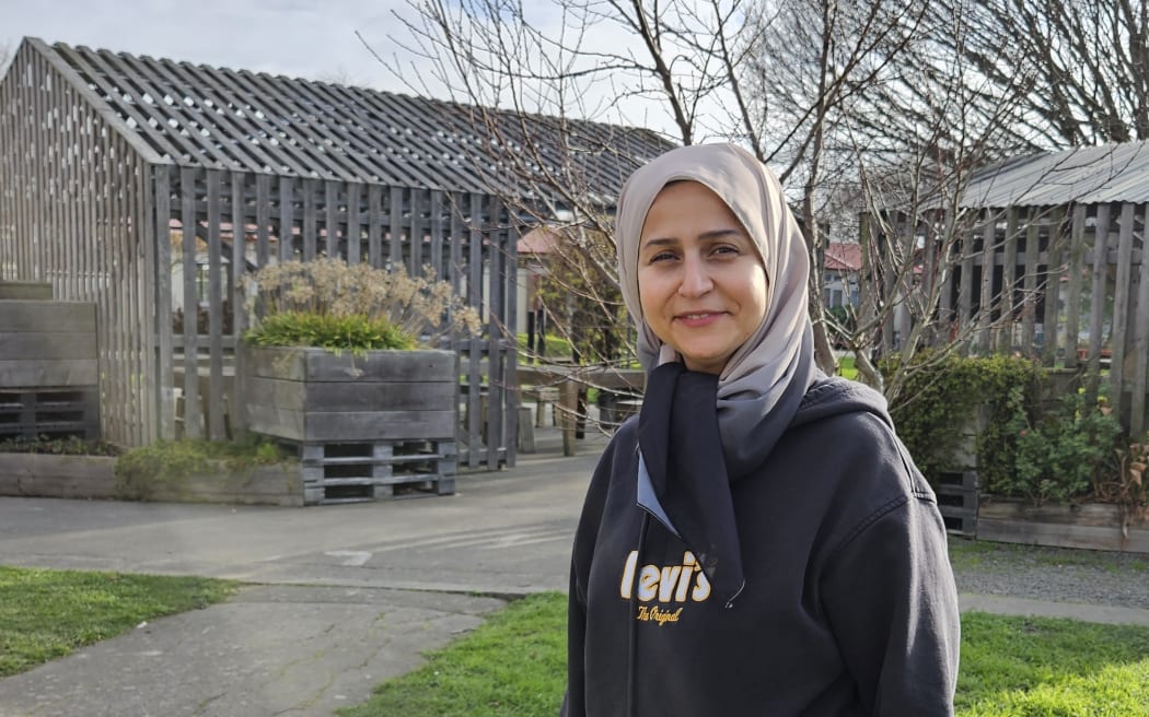 A photo of a smiling young woman wearing a head scarf standing in front of a community garden and greenhouse. The sun is shining and there is an interesting lighting effect that glances across the photo and makes the green of the grass particularly vivid.