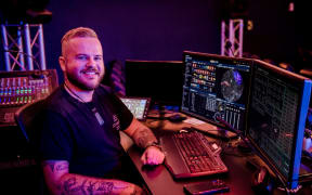Josh smiles at the camera. He sits at a control room table with models of the sky on the screens, surrounded by audiovisual equipment.