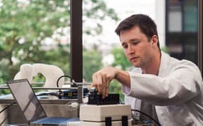 Peter operates a machine in a laboratory. He wears a white lab coat.