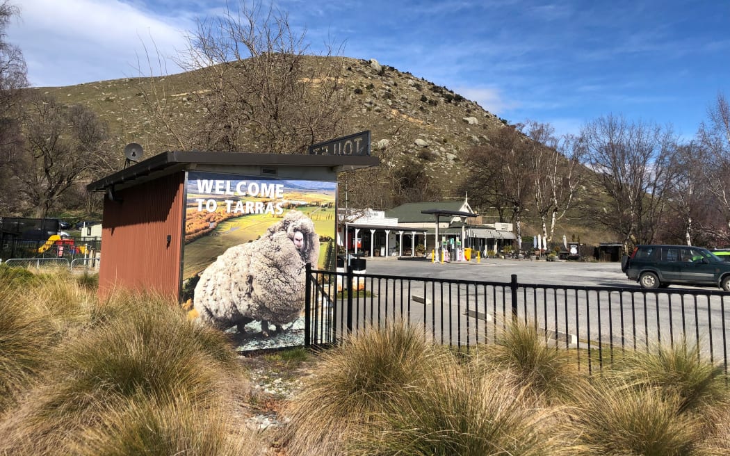 A picture of the Tarras township. In the background are small buildings and behind them, hills. The side of a public bathroom is painted with a large sheep and has text reading "WELCOME TO TARRAS".