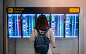 Young woman traveler with backpack and luggage looking at boarding time at departure board at terminal in international airport