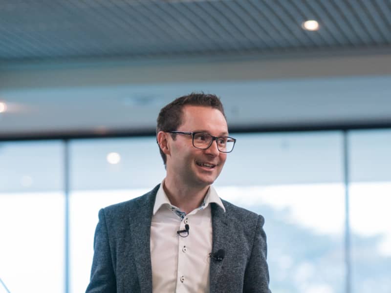James Laughlin, wearing a white shirt, grey suit jacket and glasses, makes a hand gesture while giving a presentation.
