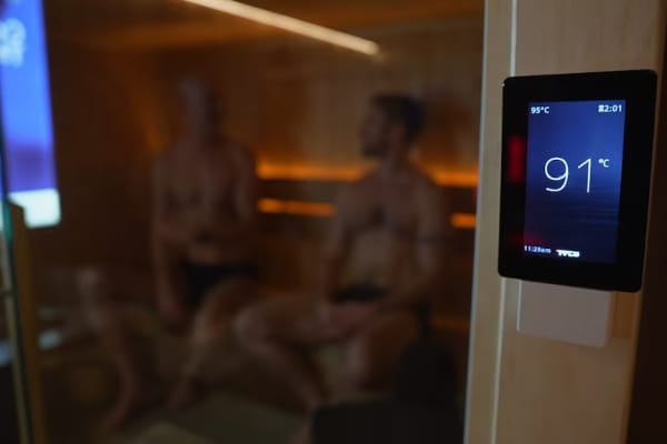 Two men sitting inside a sauna, with a thermostat at the front showing 91 degrees.