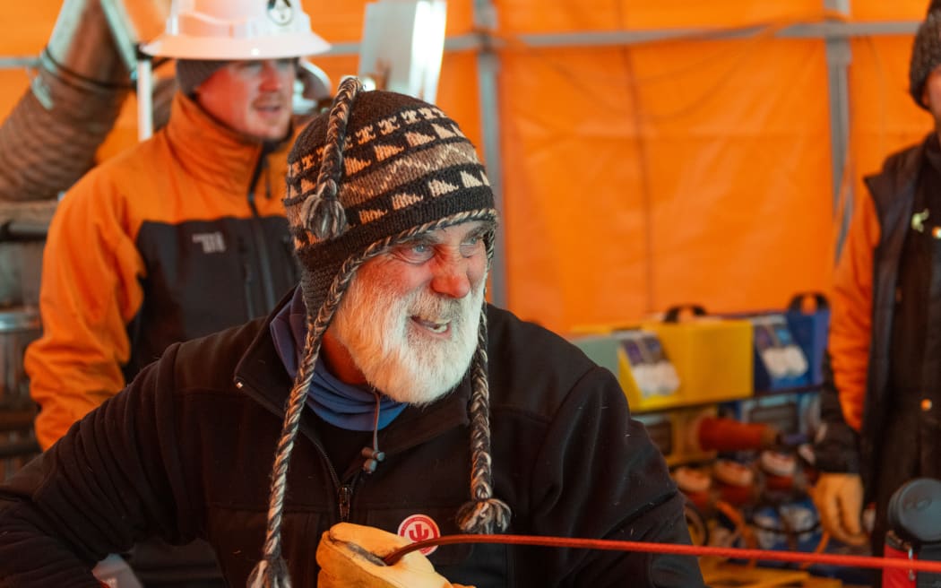 A man with a beard wearing a beanie and gloves grins while pulling on a metal rod. He is inside an orange tent and there is a man standing behind him wearing an orange and black jacket and a white hard hat.