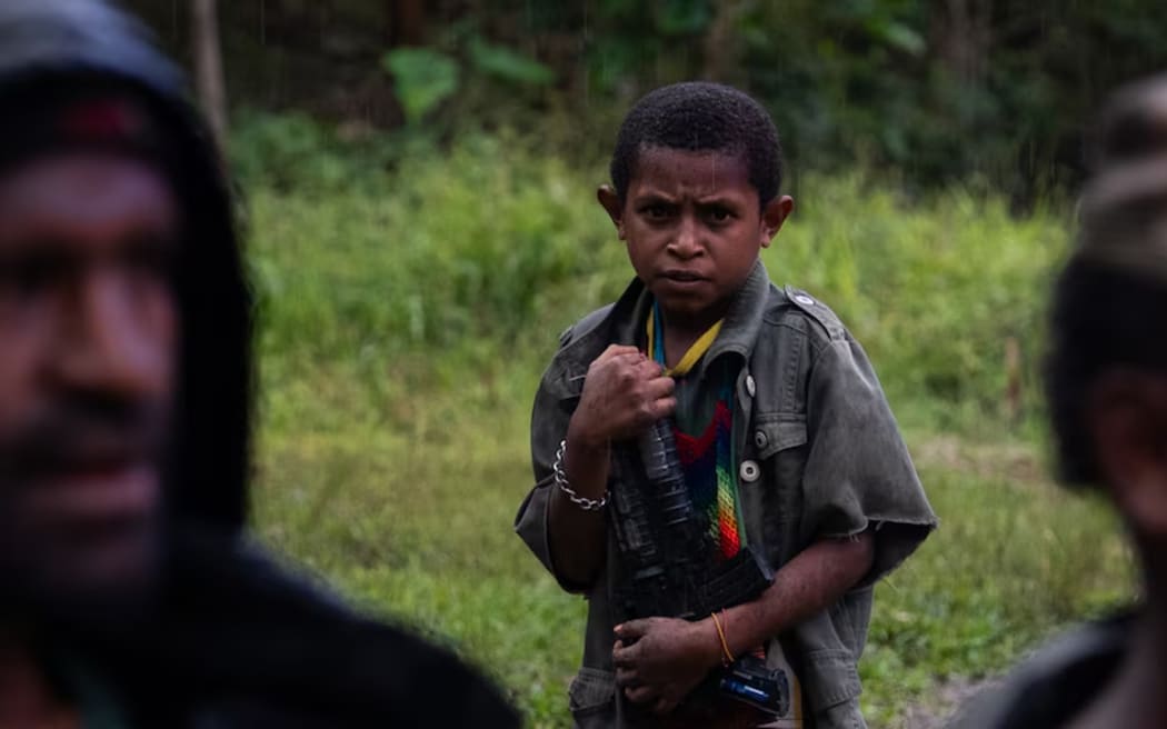 A young child holds a toy gun while the men around him wield high-powered assault rifles.