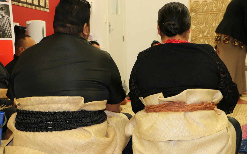 A back view of a seated Tongan man and woman in traditional dress, with mats wrapped around their waists.