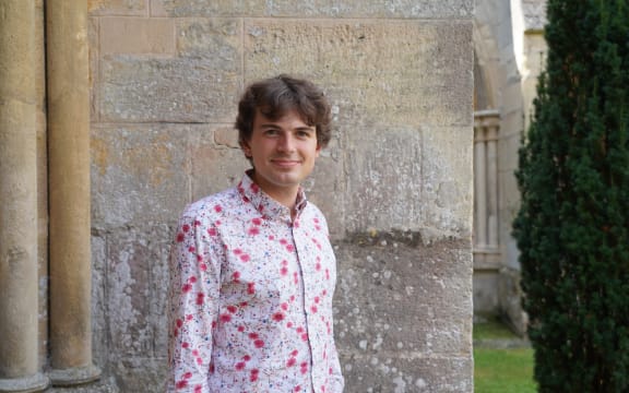 Dunedin musician Nathaniel Otley stands in front of the stone exterior of a building. He is wearing a shirt with pink with white flowers on it.