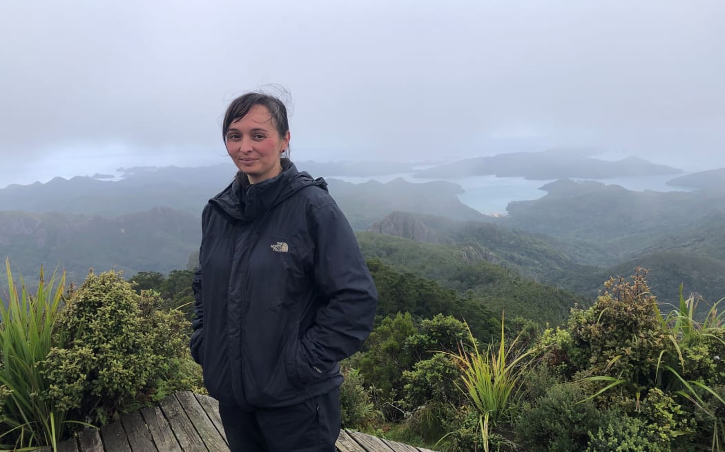 A woman in wet weather gear stands on top of a maunga on Aotea Great Barrier Island. It is cloudy.