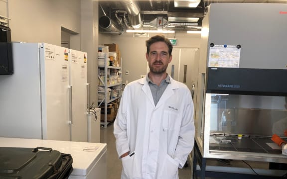 Damien wears a white lab coat. He is standing in a lab surrounded by machines, shelves and special storage units.