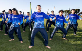 A group of teenagers in a blue sports uniform perform a haka holding hockey sticks.