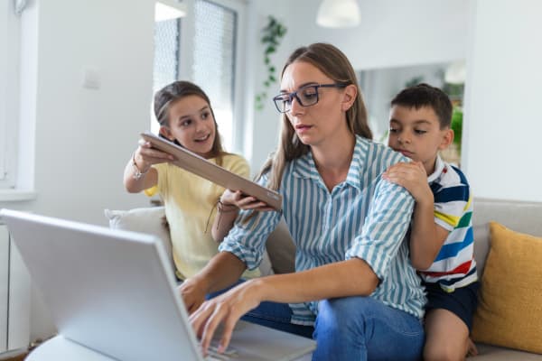 A woman working on her laptop in a living room, with kids by her side on the couch wanting her attention.