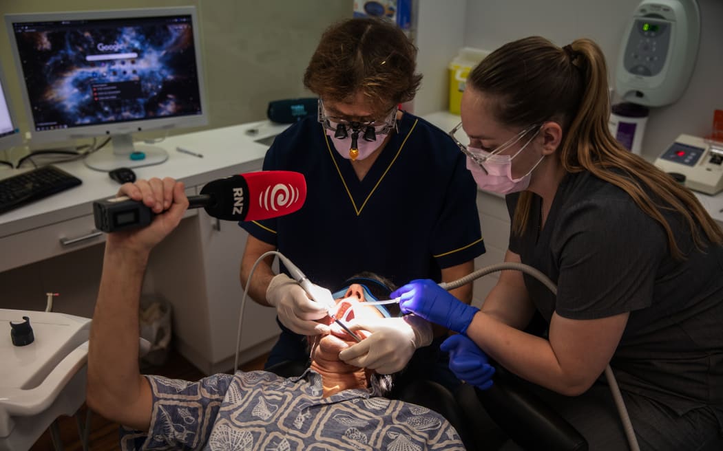 A man is lying in the dentist chair, holding a microphone in the air. The dentist and his assistant are carrying out a dental check-up.