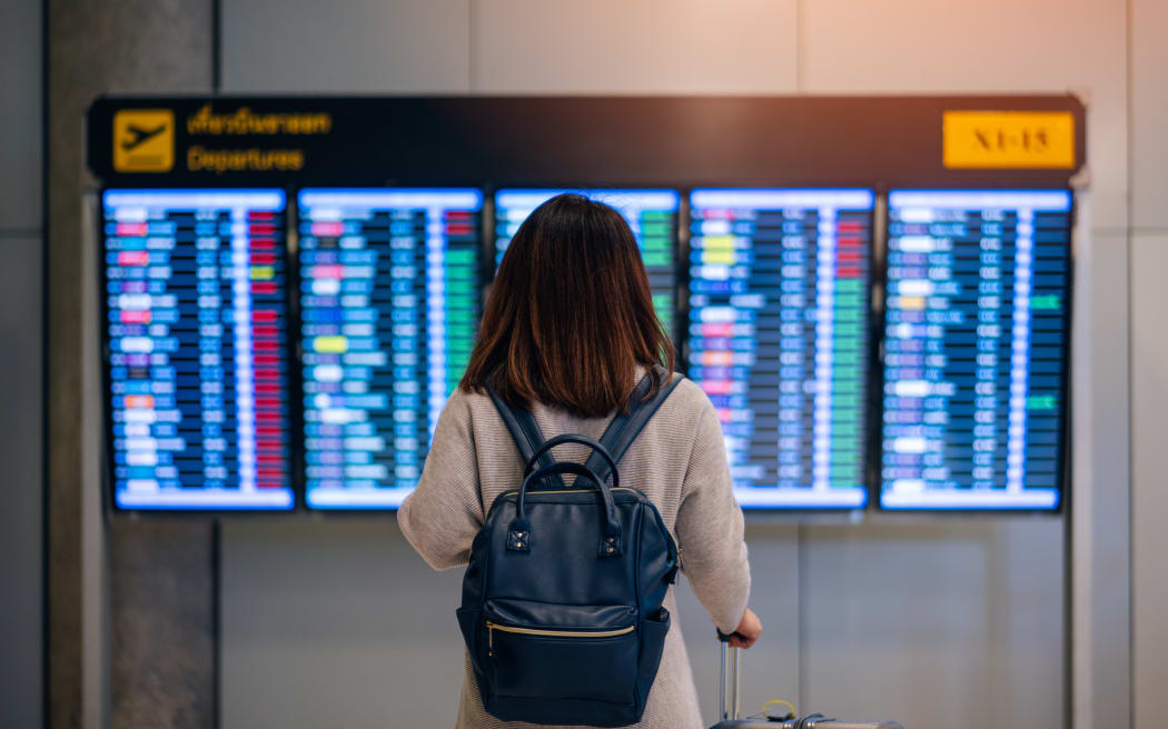 Young woman traveler with backpack and luggage looking at boarding time at departure board at terminal in international airport