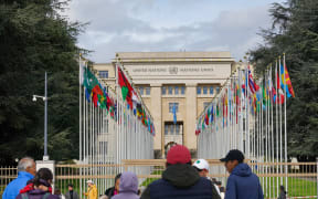People stand outside the gates of the United Nations building in Geneva, Switzerland, where the flags of member states line the entrance, symbolizing global unity. On April 1, 2024, a group of visitors stands outside the gates of the United Nations building in Geneva, Switzerland. This historic site, known as the Palais des Nations, serves as a hub for international diplomacy and peacekeeping efforts. The scene is framed by rows of flagpoles displaying the colorful flags of UN member states, emphasizing the organization's mission of fostering cooperation and unity among nations. The building's facade bears the inscription ''United Nations'' in English and French, reflecting its bilingual legacy. (Photo by Michael Nguyen/NurPhoto) (Photo by Michael Nguyen / NurPhoto via AFP)