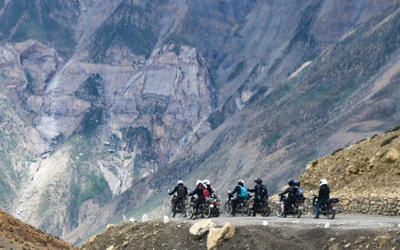 A group of motorcycle riders are on a small road halfway up a mountain. The mountain towers behind them.