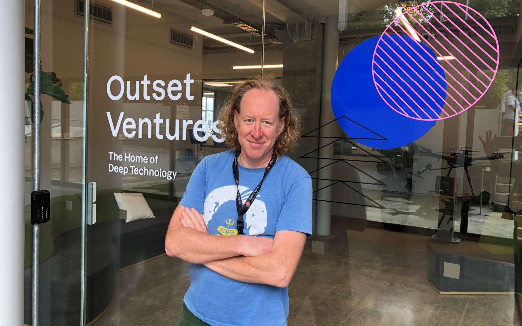 Sean smiles at the camera. He is standing outside an office building. The window of the business says "OUTSET VENTURES - THE HOME OF DEEP TECHNOLOGY".