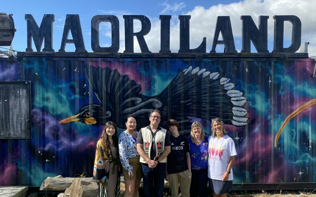 A group of people pose for a photo beneath the "MAORILAND" sign.