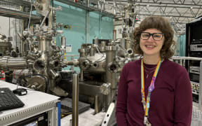 A woman with glasses and a rainbow lanyard standing in front of complicated metal machinery.