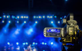 A video camera, in focus, is filming a live performance with stage lights in the distance.