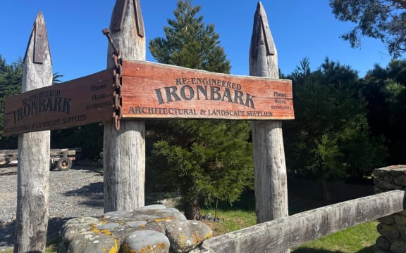 The entryway to Re-Engineered Ironbark, showing four large upright wooden poles capped with steel with a large wooden sign. In the foreground, a gateway made of riverstones