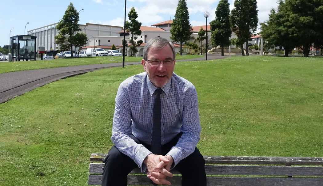 Paul Spoonley sits on the back of a bench with green grass of uni behind